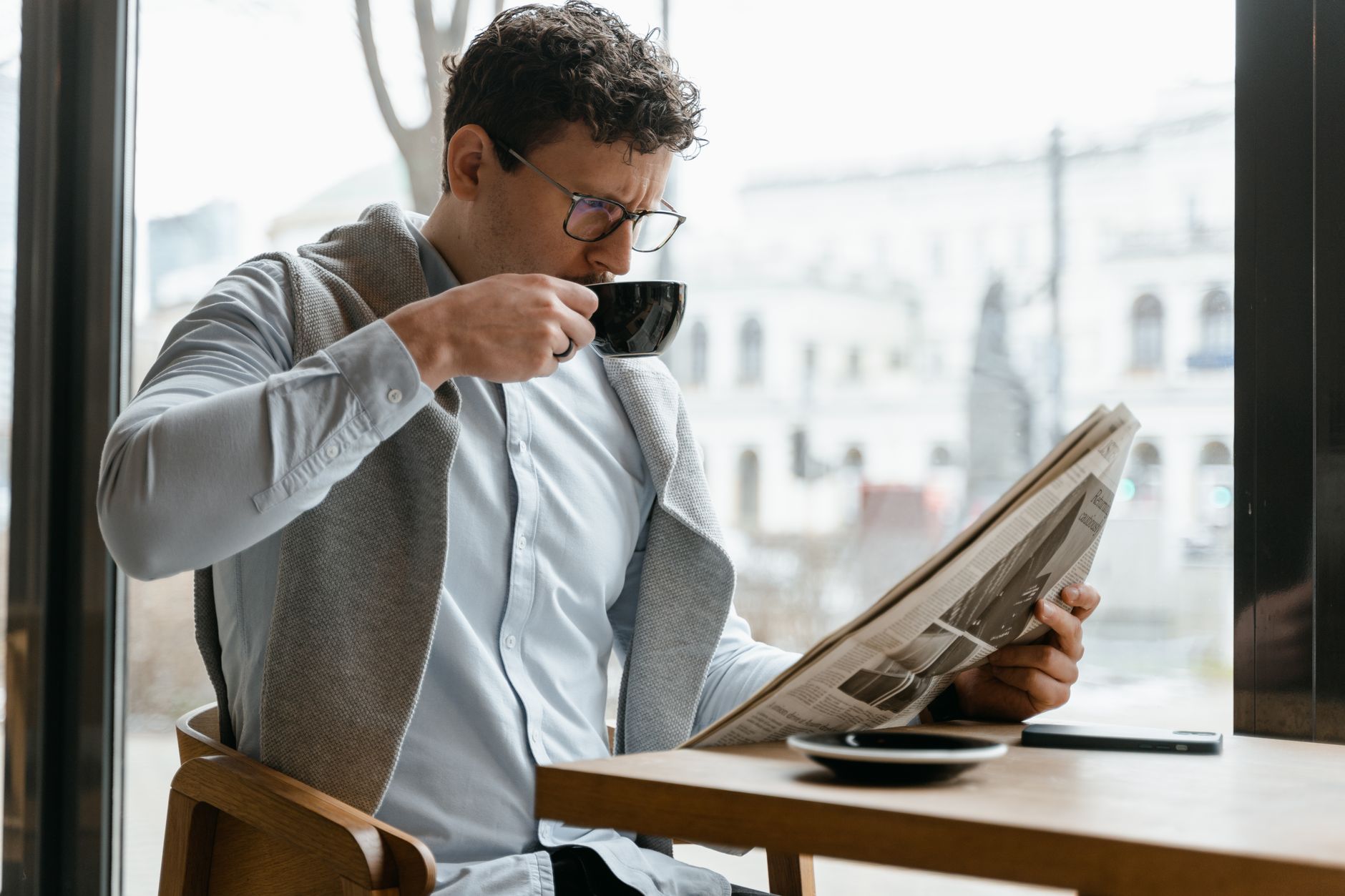 Adult man wearing eyeglasses sipping coffee and reading a newspaper in a cozy cafe setting. — Sensible Medical insurance Prep